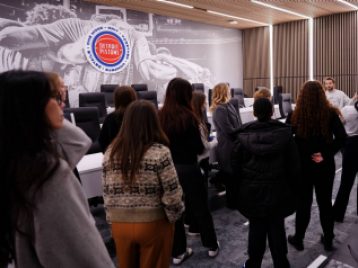 Guide at the Detroit Pistons giving a tour of a sound proof meeting room with students gathered together and the Pistons' logo on the wall.